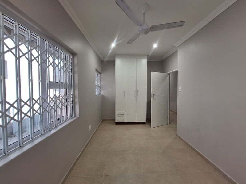 A clean, empty bedroom featuring neutral tiled flooring, a built-in wardrobe, a ceiling fan, and security gates on the windows.