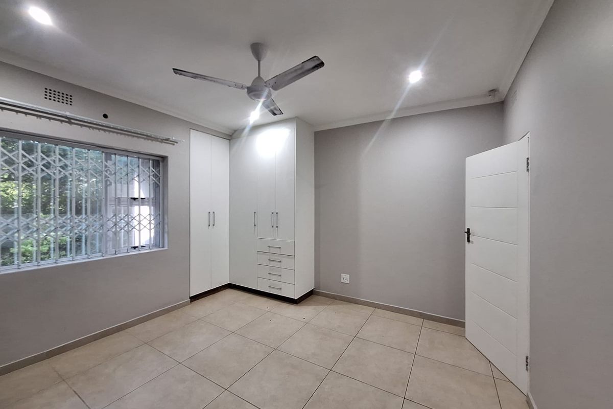 A clean, neutral bedroom featuring tiled flooring, built-in cupboards, a ceiling fan, and secure window treatments, offering a bright and versatile living space.