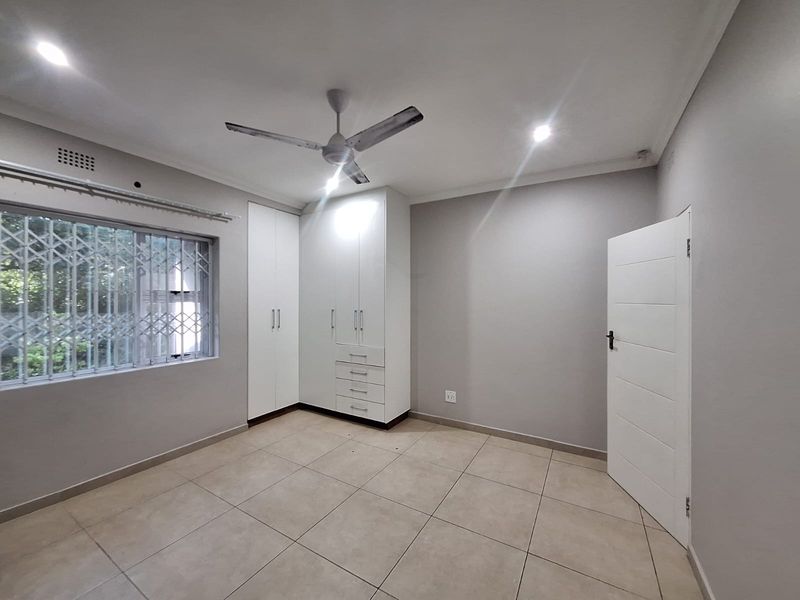 A clean, neutral bedroom featuring tiled flooring, built-in cupboards, a ceiling fan, and secure window treatments, offering a bright and versatile living space.