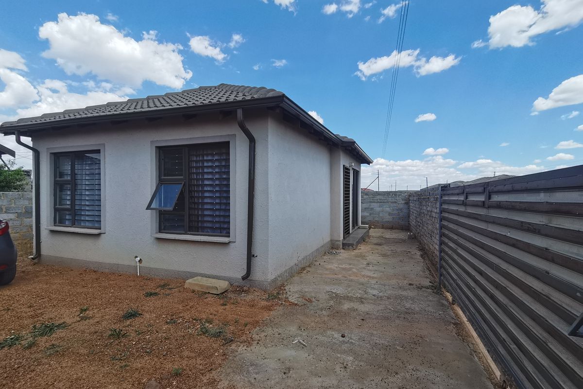 Rear view of the property featuring a tiled roof, aluminium window frames, and a concrete apron area enclosed by a boundary wall and metal fencing.
