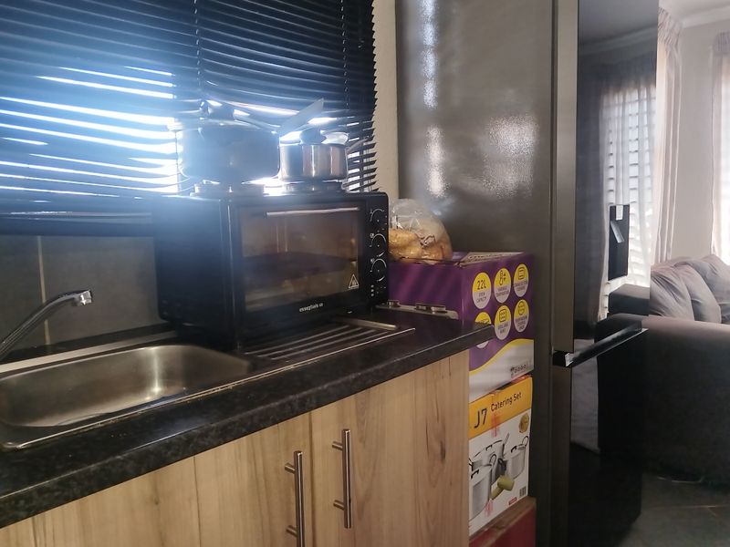 A compact kitchen area featuring laminate cabinetry, a stainless steel sink, and dark countertops, with venetian blinds providing natural light control.