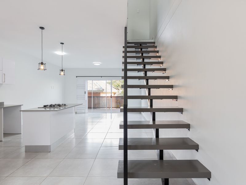 Modern open-plan living area featuring a sleek kitchen with island, gas cooktop, and a striking floating staircase leading to the upper level.