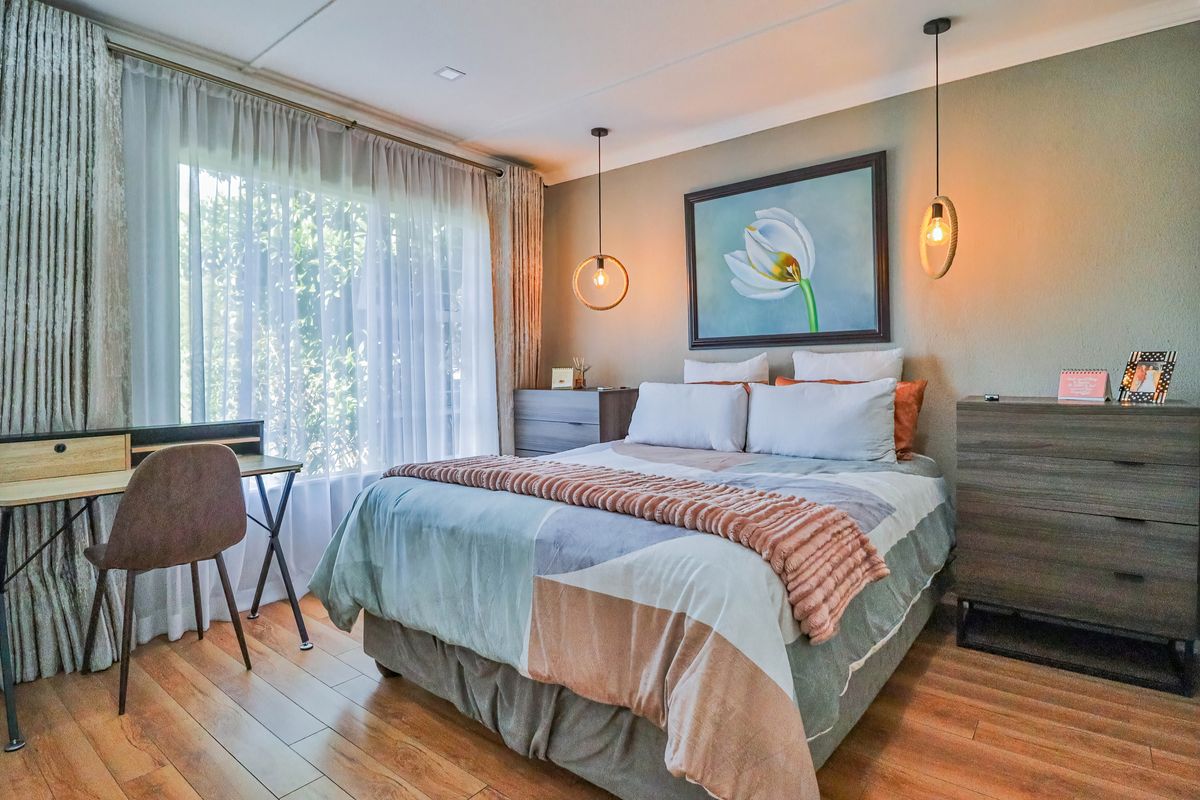 A well-maintained bedroom featuring laminate flooring, modern pendant lighting, and large windows with sheer curtains, creating a bright and comfortable living space.