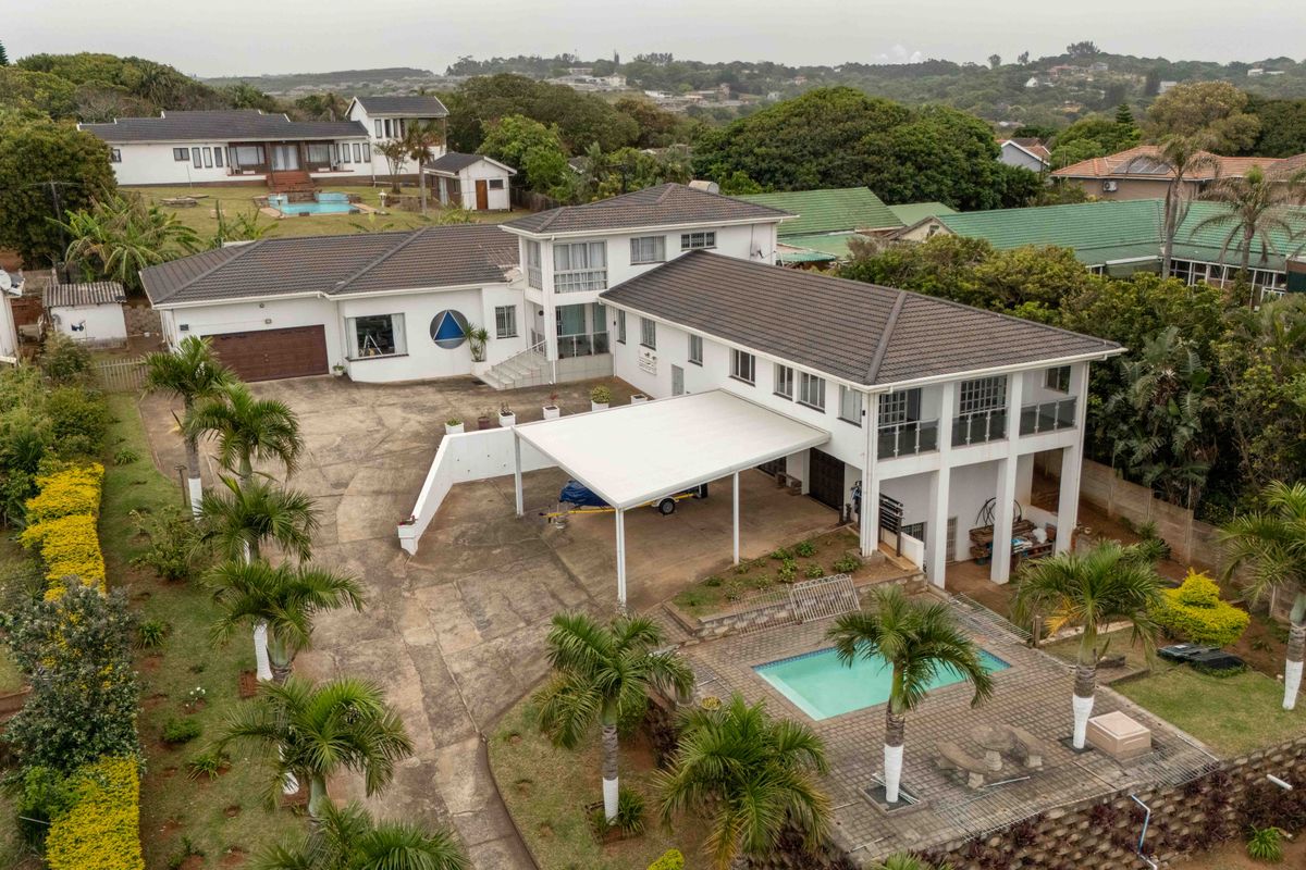 Aerial view of a multi-level residential property featuring a swimming pool, large paved driveway, carport, and balcony overlooking the surrounding landscape.