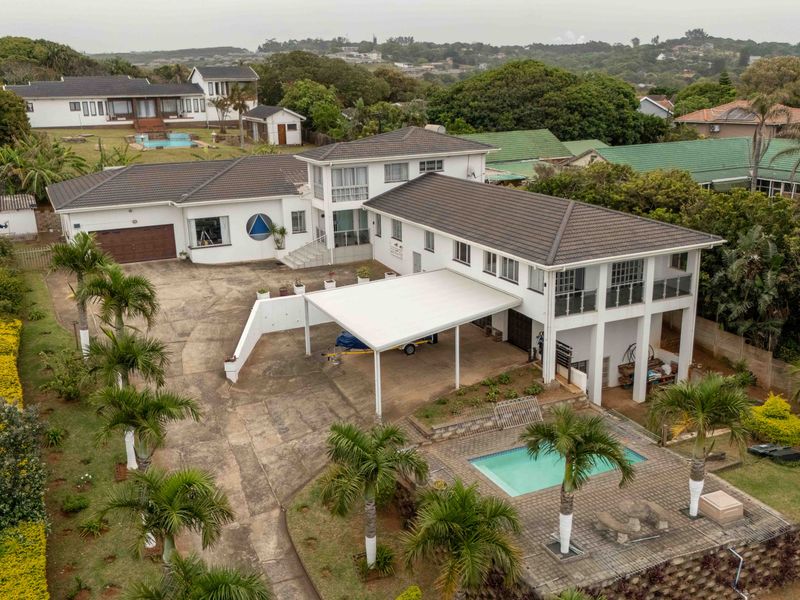 Aerial view of a multi-level residential property featuring a swimming pool, large paved driveway, carport, and balcony overlooking the surrounding landscape.