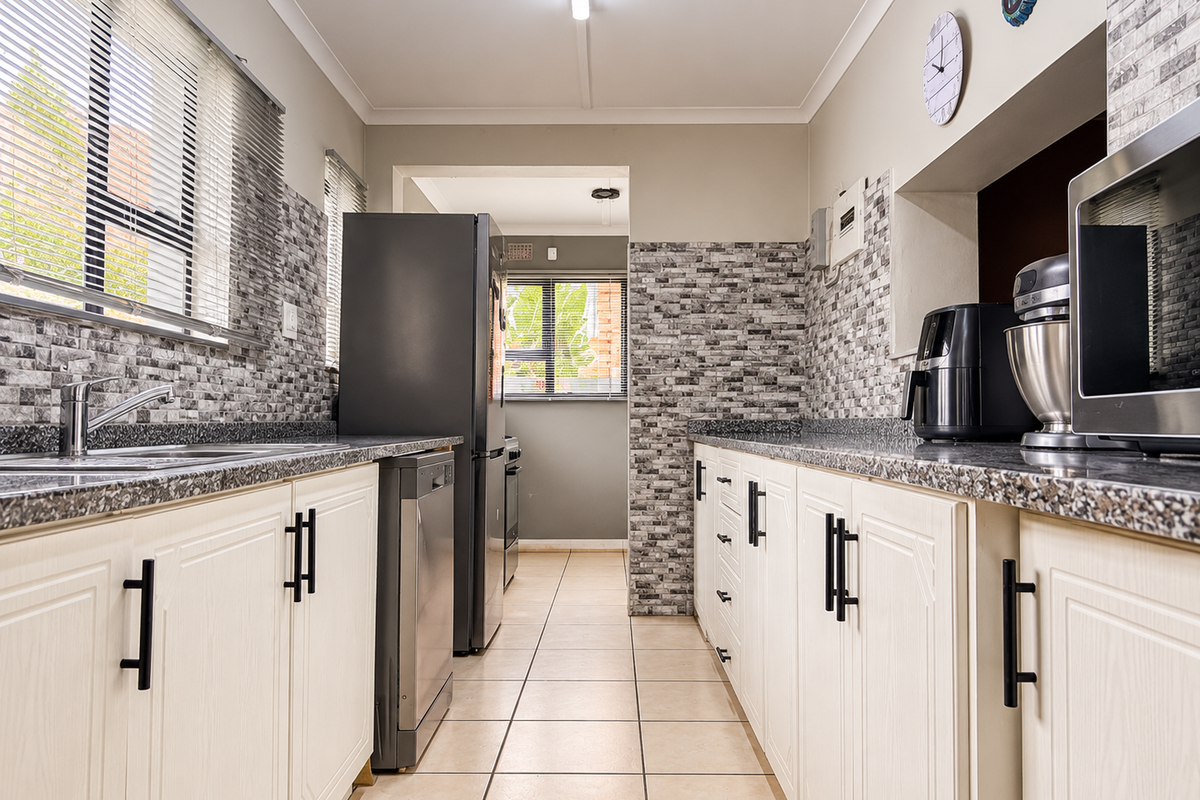 A functional galley-style kitchen featuring granite countertops, tiled backsplash, ample cabinetry, and tiled flooring with natural light from large windows.
