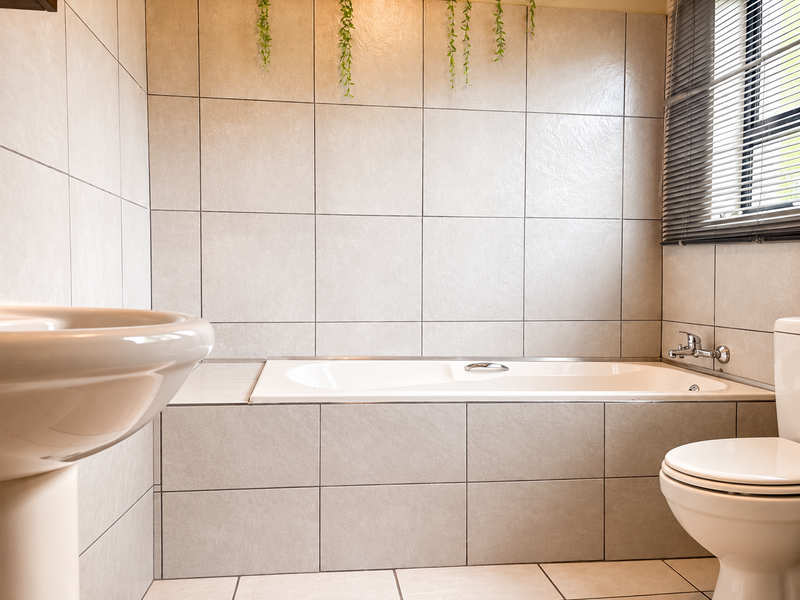 A clean, neutral-toned bathroom featuring a built-in bathtub, pedestal basin, and toilet, finished with floor-to-ceiling ceramic tiling and functional window blinds.