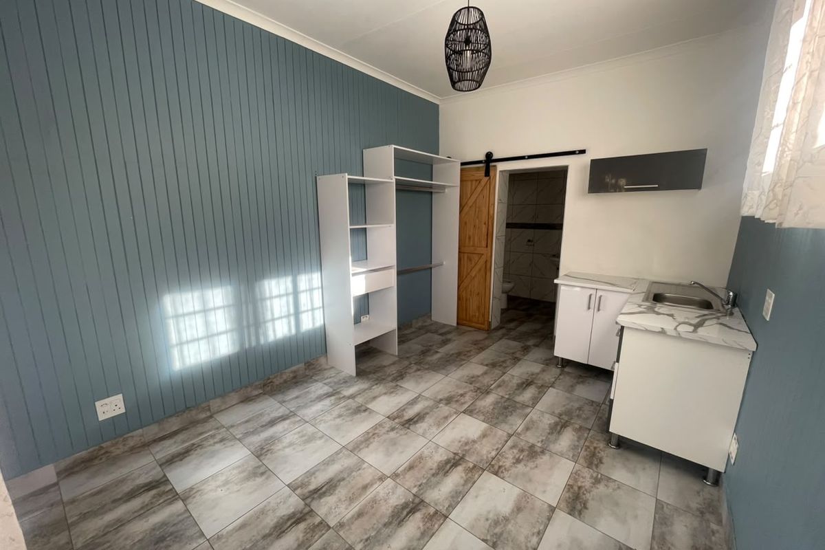 Open-plan studio space featuring tiled flooring, a panelled feature wall, built-in shelving, and a compact kitchenette with sink and cabinetry.