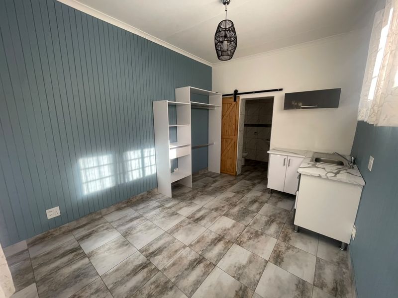 Open-plan studio space featuring tiled flooring, a panelled feature wall, built-in shelving, and a compact kitchenette with sink and cabinetry.