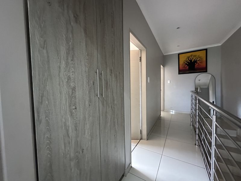 A bright, modern hallway featuring light wood-finish built-in cupboards, large format floor tiles, and a sleek stainless steel balustrade overlooking the staircase.