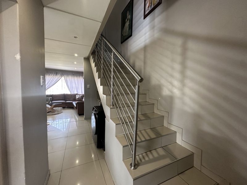 A bright hallway featuring a tiled staircase with a modern stainless steel balustrade, leading towards an open-plan living area with polished floor tiles.