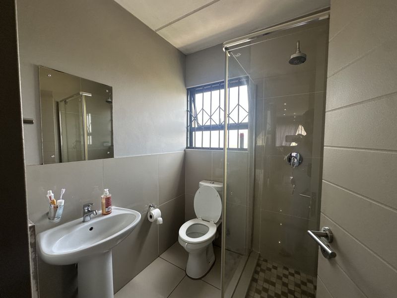 A functional bathroom featuring a pedestal basin, toilet, and a glass-enclosed shower with tiled finishes and security bars on the window.