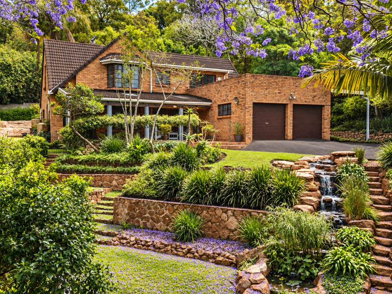 A beautiful double-story brick house featuring a double garage, tiered stone retaining walls, and a picturesque landscaped garden with a built-in water feature.