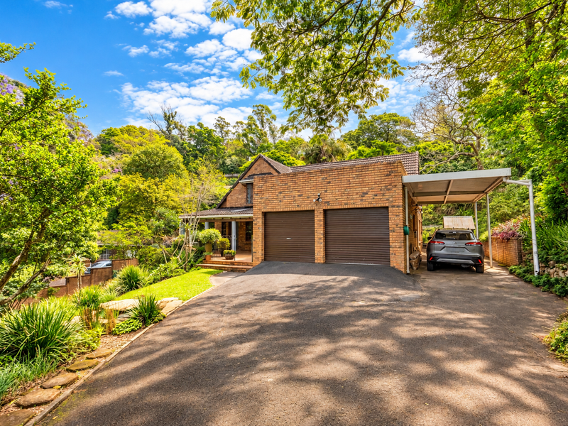 Front view of a brick house featuring a double garage, an attached carport, and a paved driveway surrounded by mature trees and landscaped garden.