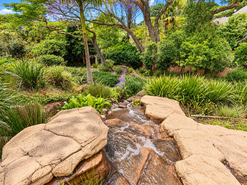 A lush, well-maintained garden featuring a tiered rock water feature surrounded by mature trees and diverse plantings.