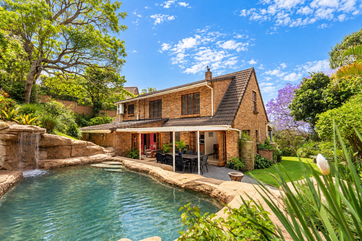 Rear view of a brick house featuring a landscaped swimming pool with a rock waterfall, a covered patio area, and lush garden surroundings.