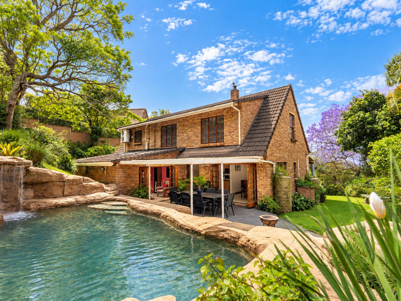 Rear view of a brick house featuring a landscaped swimming pool with a rock waterfall, a covered patio area, and lush garden surroundings.