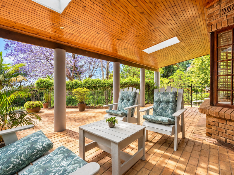 A covered outdoor patio featuring a timber-clad ceiling with skylights, brick paving, and structural columns, overlooking a lush garden area.