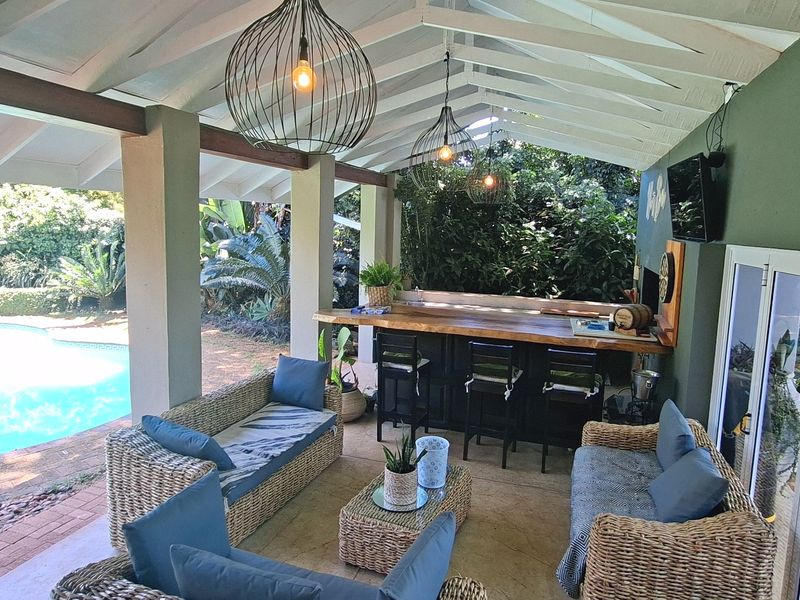 Covered outdoor patio featuring a built-in bar, braai area, and exposed timber beam ceiling, overlooking the swimming pool and garden.