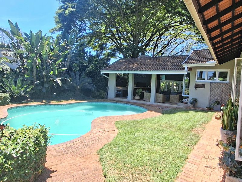 A well-maintained outdoor pool area featuring a swimming pool, brick paved surround, and a covered patio area set within a lush garden.