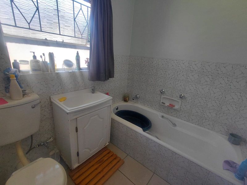A functional bathroom featuring a built-in bathtub, vanity cabinet with sink, and toilet, finished with dated wall tiling and security bars on the window.