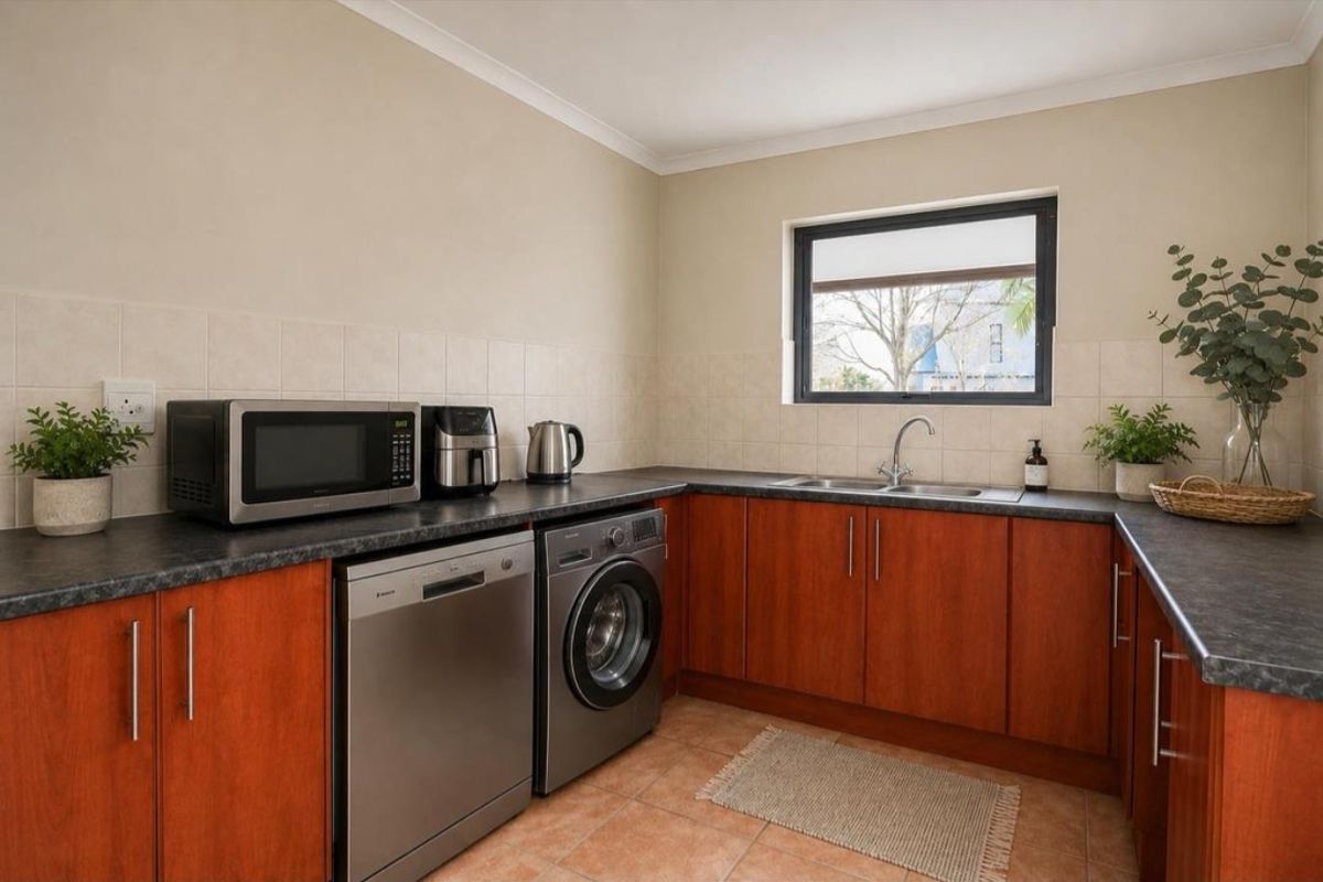 A functional kitchen featuring wood-finish cabinetry, dark laminate countertops, a double stainless steel sink, and tiled flooring with a neutral backsplash.