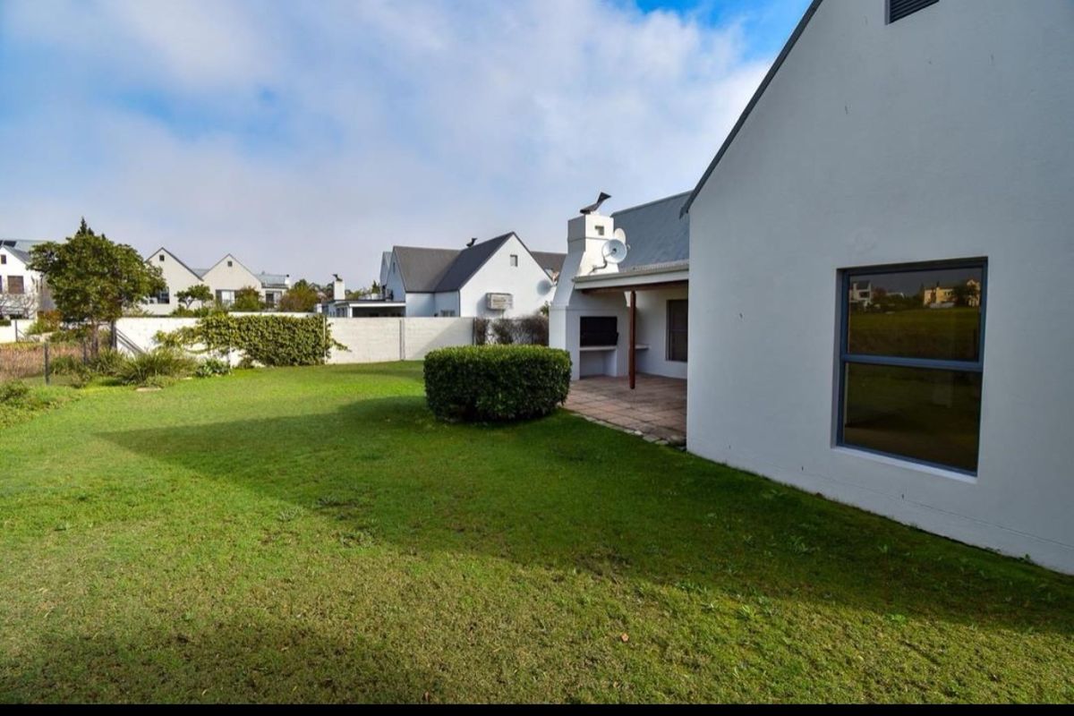 Rear view of the property featuring a well-maintained lawn, a covered patio area with a built-in braai, and secure boundary walling.