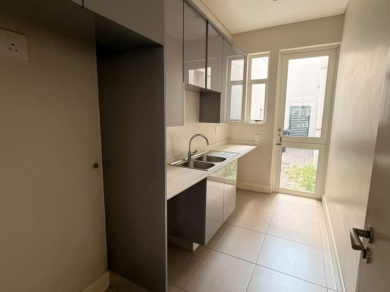 A clean, modern scullery featuring grey built-in cabinetry, a stainless steel double sink, tiled flooring, and direct access to the exterior.