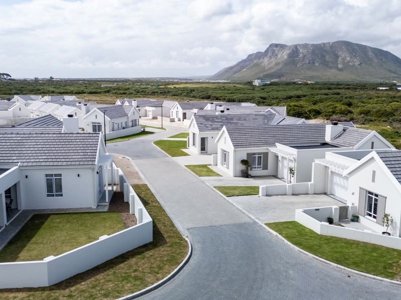 Aerial view of a modern residential estate featuring uniform white houses with grey tiled roofs, paved driveways, and manicured lawns against a mountain backdrop.