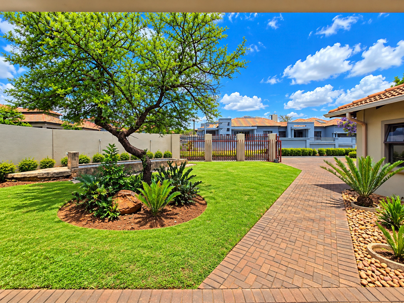 A well-maintained landscaped garden featuring a paved walkway, mature tree, and secure boundary walling with a metal gate.