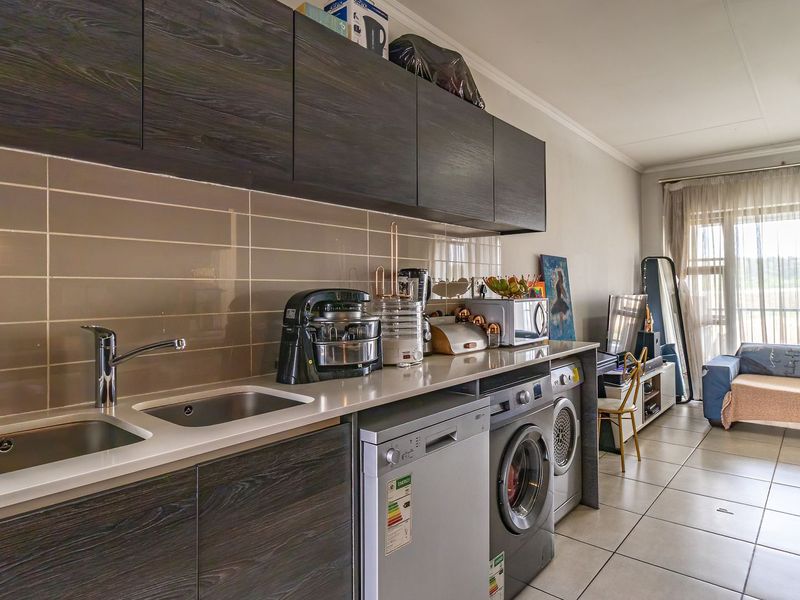 Modern open-plan kitchen and living area featuring dark wood cabinetry, integrated appliances, tiled backsplash, and neutral ceramic flooring leading to a balcony.