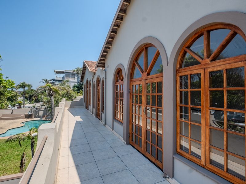 Spacious upper-level balcony featuring arched wooden french doors, tiled flooring, and a parapet wall overlooking the garden and pool area.