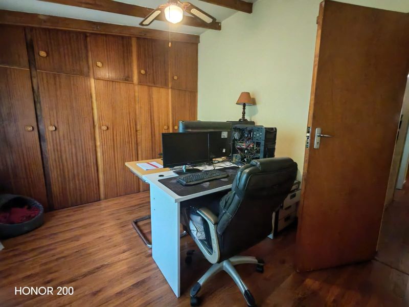 A bedroom featuring built-in wooden cupboards, exposed timber ceiling beams, and laminate flooring, currently utilized as a home office space.