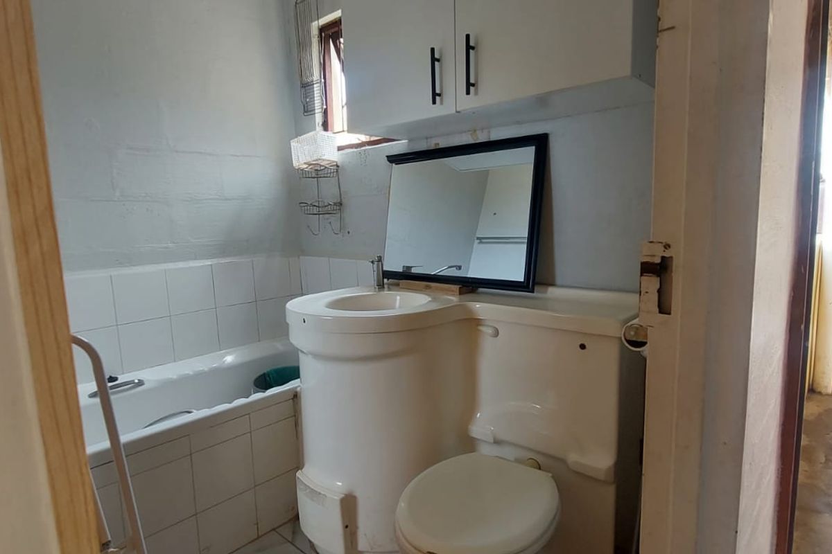 A compact bathroom featuring a combined basin and toilet unit, a bathtub with tiled splashback, and overhead storage cabinetry.