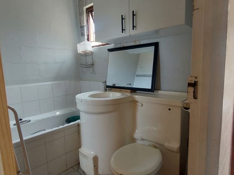 A compact bathroom featuring a combined basin and toilet unit, a bathtub with tiled splashback, and overhead storage cabinetry.