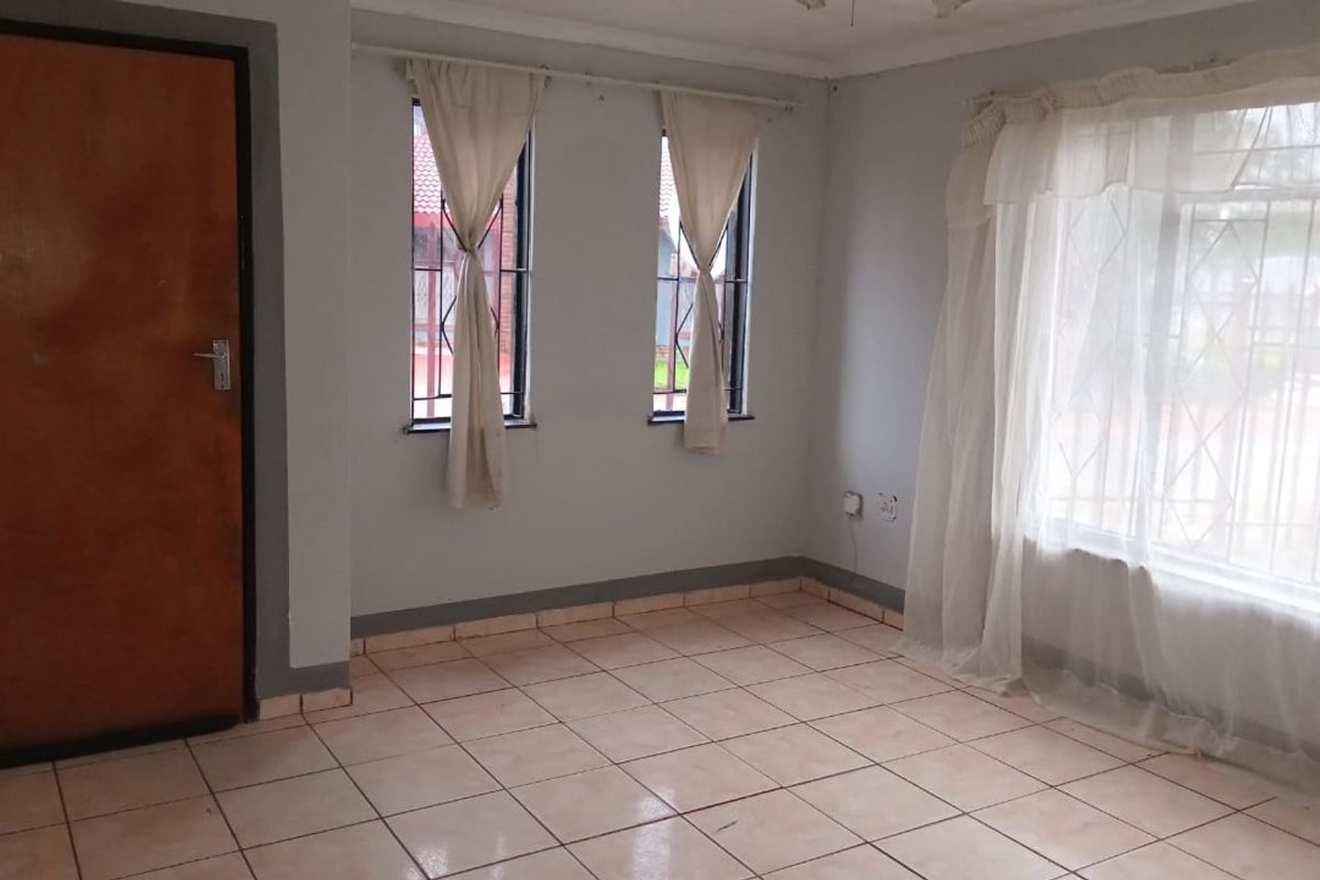 An empty living room featuring tiled flooring, large windows with security bars, and a wooden door, offering a blank canvas for interior design.
