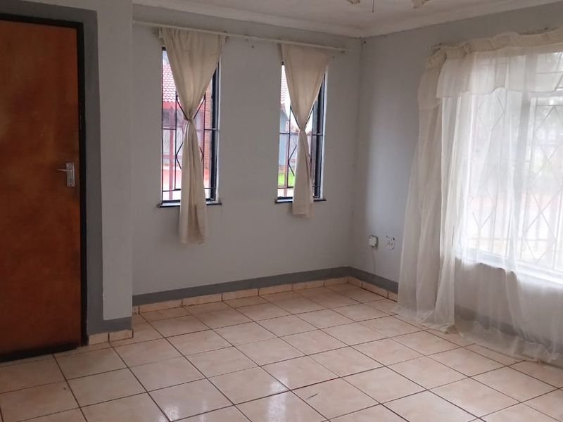An empty living room featuring tiled flooring, large windows with security bars, and a wooden door, offering a blank canvas for interior design.