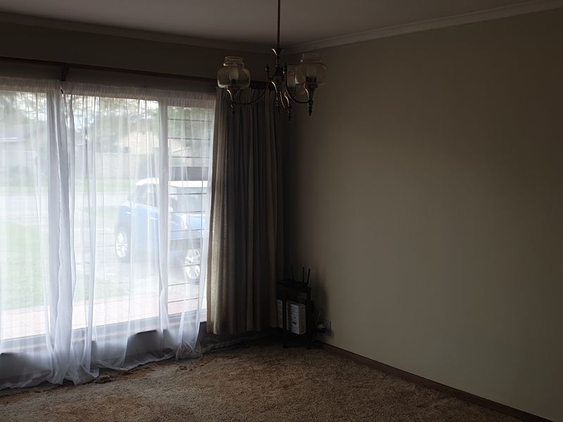 A spacious, empty living room featuring large windows that provide natural light, a classic ceiling chandelier, and carpeted flooring.