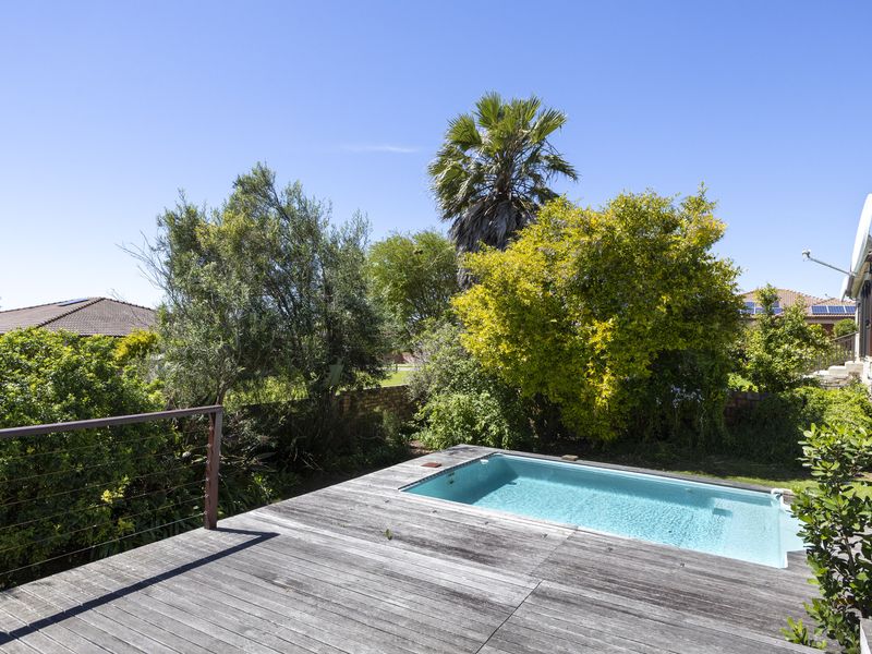 Outdoor pool area featuring a swimming pool set within a timber deck, surrounded by established garden landscaping and a brick boundary wall.
