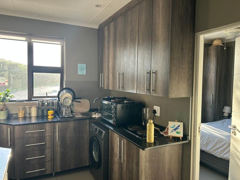 Compact kitchen featuring modern wood-grain cabinetry, dark granite countertops, integrated hob, and built-in oven, with a view into the adjacent bedroom.