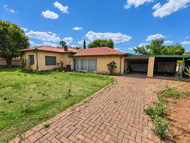 Rear view of a single-story house featuring a corrugated metal roof, brick driveway, and an attached carport, set within a large garden area.