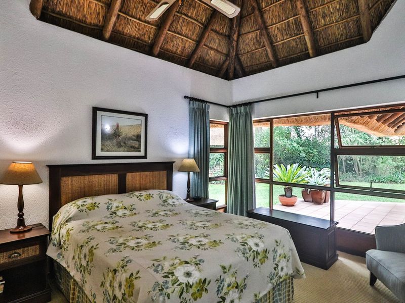 A cozy bedroom featuring a unique thatched ceiling, ceiling fan, and large windows providing natural light and views of the garden patio.