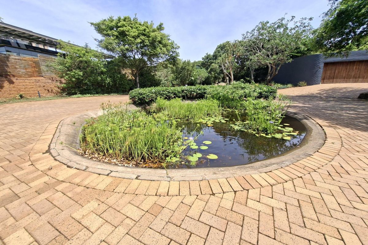 A well-maintained circular water feature set within a brick-paved driveway area, surrounded by established garden landscaping and mature trees.
