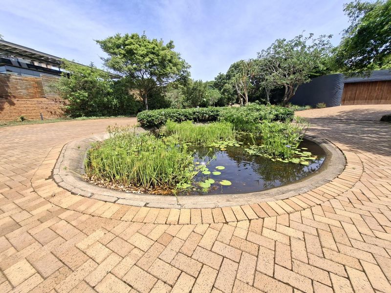 A well-maintained circular water feature set within a brick-paved driveway area, surrounded by established garden landscaping and mature trees.