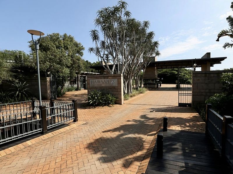 The entrance to Hawaan Forest Estate featuring a paved driveway, security gate, and stone wall signage surrounded by mature trees.