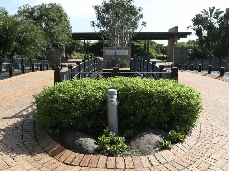 A well-maintained residential estate entrance featuring a brick-paved driveway, pedestrian bridge, and landscaped garden area with integrated lighting.