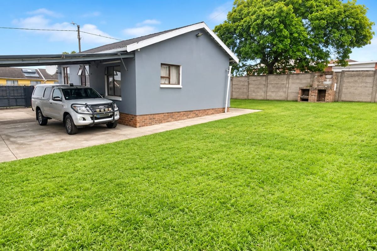 Spacious rear garden featuring a large lawn, a covered carport, and a built-in brick braai area, enclosed by precast concrete boundary walls.