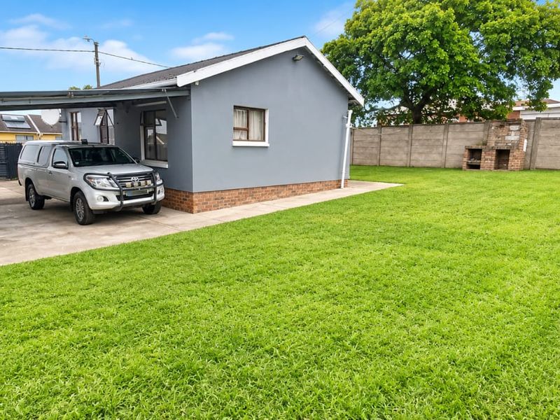 Spacious rear garden featuring a large lawn, a covered carport, and a built-in brick braai area, enclosed by precast concrete boundary walls.