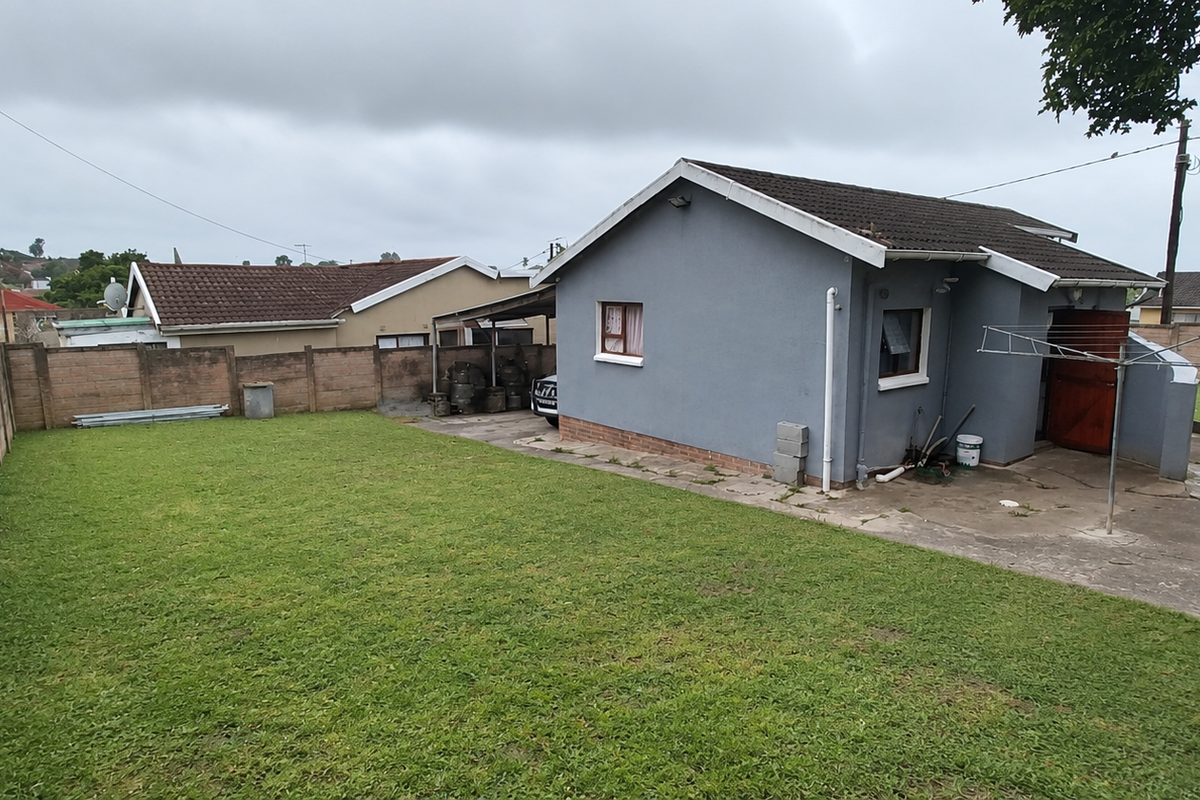Rear view of the property featuring a spacious lawn, concrete patio area, and perimeter boundary walls under a tiled roof structure.