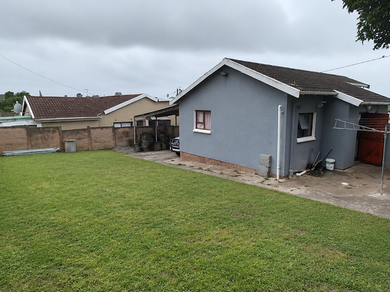 Rear view of the property featuring a spacious lawn, concrete patio area, and perimeter boundary walls under a tiled roof structure.